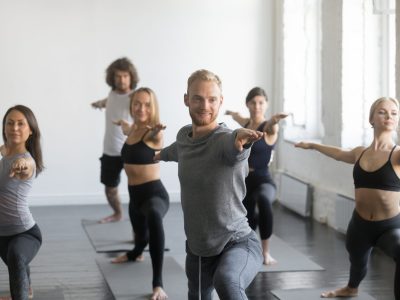 Group of young sporty smiling people practicing yoga lesson with instructor, standing in Warrior Two exercise, Virabhadrasana 2 pose, working out, indoor close up image, studio. Wellbeing concept